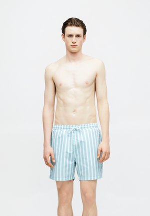 Jeune homme à la peau claire et aux cheveux mouillés, debout, portant un short de bain bleu clair à rayures verticales blanches avec un cordon de serrage.