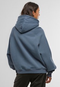 Nadmerná modrá mikina s kapucňou, textúrovaným povrchom, veľkou kapucňou a rebrovanými manžetami a lemom. Mäkká látka a voľný strih.