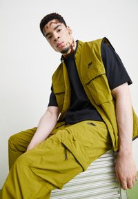 Casaquinho sem mangas em verde-oliva com bolsos, por cima de uma t-shirt preta, combinado com calças a condizer com fechos de correr e detalhes pretos.