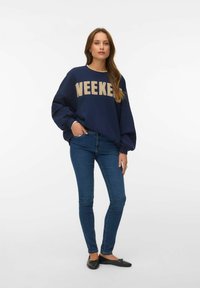 Femme portant un sweat-shirt ample bleu marine avec le texte "WEEKEND", un jean slim bleu et des ballerines noires, debout devant un fond blanc uni.
