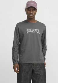 Graues Langarmshirt mit einem weißen "WORLD TOUR"-Grafikdesign, kombiniert mit dunklen Jeans und einer helllila Mütze.