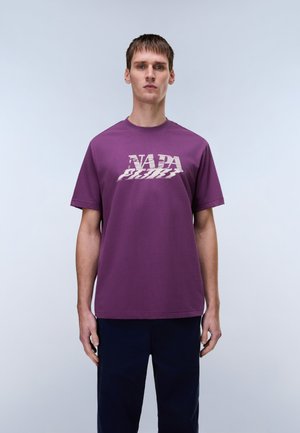 Mann trägt ein lila T-Shirt mit weißem, verzerrtem "NAPAPIJRI"-Schriftzug, steht vor einem schlichten hellgrauen Hintergrund.