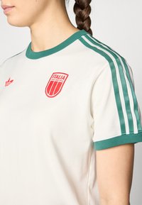 Cream-colored short-sleeve t-shirt with green ribbed collar and stripes. Features a red "Italia" badge on the chest and a small Adidas logo.