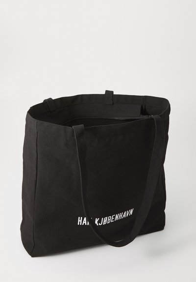 Geantă din pânză neagră cu două mânere lungi, având un logo alb pe față și un vârf deschis. Textură netedă, formă rectangulară.
