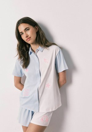 Jonge vrouw leunt tegen een witte muur, draagt een pastelblauw en wit kortärmelig shirt en bijpassende shorts met roze zeedierprints.