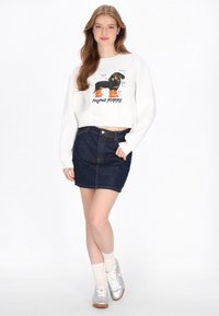 Sudadera blanca corta con un gráfico de un perro caricaturesco; combinada con una falda vaquera azul oscuro y zapatillas plateadas, acentuada con calcetines blancos.