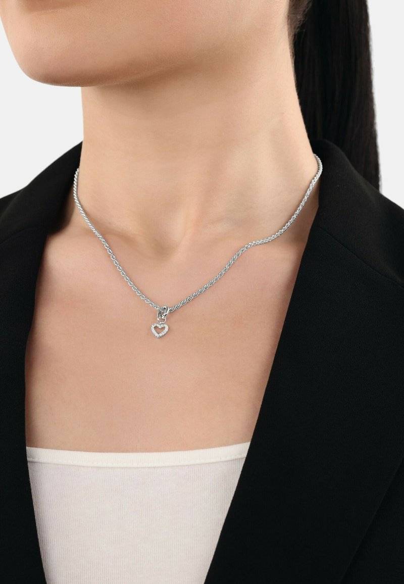 Collana in argento con un design intrecciato e un pendente a forma di cuore, decorato con piccoli cristalli trasparenti, indossata su una maglia chiara.