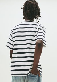 Persoon met dreadlocks die een wit en zwart gestreept shirt met korte mouwen draagt, met één hand in de zak van lichtblauwe jeans, met de rug naar de kijker gekeerd.