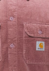 Camisa de pana rosa con acanalado vertical, dos bolsillos frontales y un pequeño parche blanco de Carhartt en el bolsillo izquierdo.