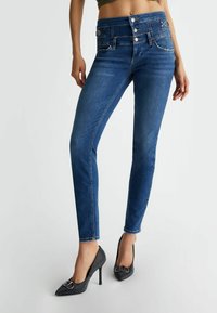 Jeans blu skinny in denim con cinque tasche, vita con più bottoni e cuciture decorative. Indossati con tacchi neri a punta.