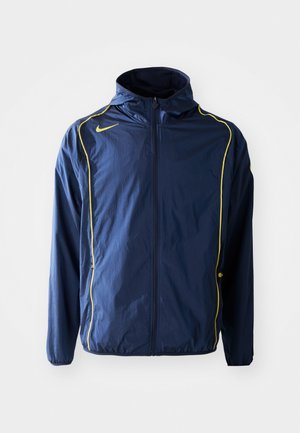 Marinblå Nike-jacka med gula detaljer, lättviktsmaterial, hel dragkedja fram, justerbar huva och elastiska muddar. Enkelt design, inga ytterligare detaljer.