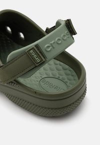 Sandale Crocs vert olive avec une semelle en caoutchouc texturée, une sangle réglable et un motif circulaire sur la semelle intérieure, comportant le logo de la marque.