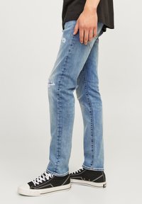 Jeans de mezclilla azul claro con un corte ajustado, que presentan elementos desgastados cerca de las rodillas, combinados con zapatillas negras y cordones blancos.