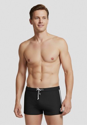 Fit aussehender Mann stehend, trägt schwarze Badeshorts mit weißen Seitenstreifen und weißem Kordelzug, vor einem schlichten hellen Hintergrund.
