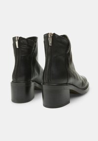 Botas de tobillo de cuero negro con textura lisa, punta redonda y tacón rectangular. Presentan cremalleras plateadas en la parte trasera.