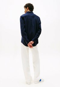 Mann mit lockigem Haar steht von hinten und trägt ein dunkelblaues Langarmhemd, weiße Hose und weiße Sneaker mit blauen Details.