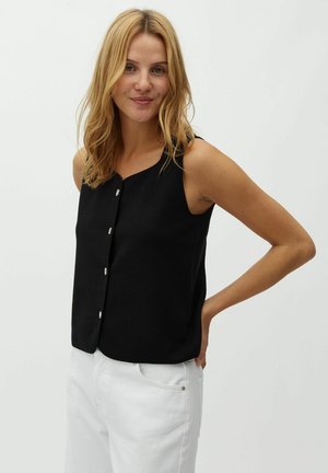 Szőke nő ujjatlan fekete gombos topban és fehér nadrágban, egy kézzel a csípőjén pózol egyszerű háttér előtt.