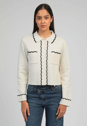 Femme portant un cardigan crème boutonné avec un galon ondulé noir sur le col, les poches et les manches, associé à un jean taille haute bleu sur fond gris.