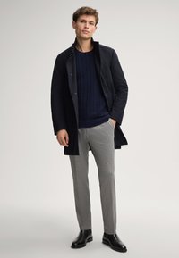 Cappotto blu navy con colletto rialzato sopra un maglione blu navy a trama. Pantaloni grigi e stivali in pelle nera completano l'outfit.