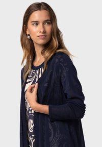 Marinblå, lätt cardigan med öppen front och långa ärmar, lager på lager över en mönstrad krämfärgad topp. Slät textur med en subtil design.