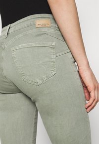 Jeans de mezclilla verde claro con un diseño ajustado, que presentan un bolsillo trasero sin costuras con costuras decorativas y un parche de cuero de la marca en la cinturilla.