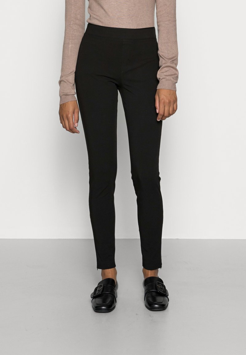 Svarta stretchbyxor med en åtsittande design, slät textur och ankel-längd, i kombination med svarta slip-on skor som har en spänndetalj.