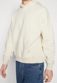 Gräddfärgad hooded sweatshirt av mjukt material, med en känguruficka och ribbade ärmslut, tillsammans med denimjeans.