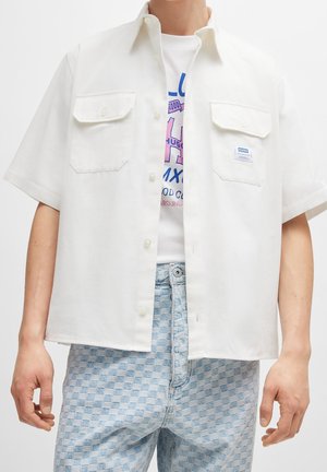 Hvid kortærmet skjorte med knapper og to brystlommer, båret åben over en hvid grafisk t-shirt, kombineret med lyseblå ternede jeans.