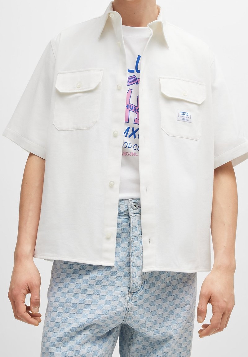 Chemise blanche à manches courtes boutonnée avec deux poches poitrine, portée ouverte sur un t-shirt blanc graphique, associée à un jean à carreaux bleu clair.