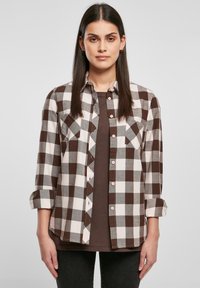 Camicia a quadri marrone e bianca con maniche lunghe, due tasche anteriori e bottoni rosa sopra una maglia marrone scuro. Tessuto morbido con un design classico.