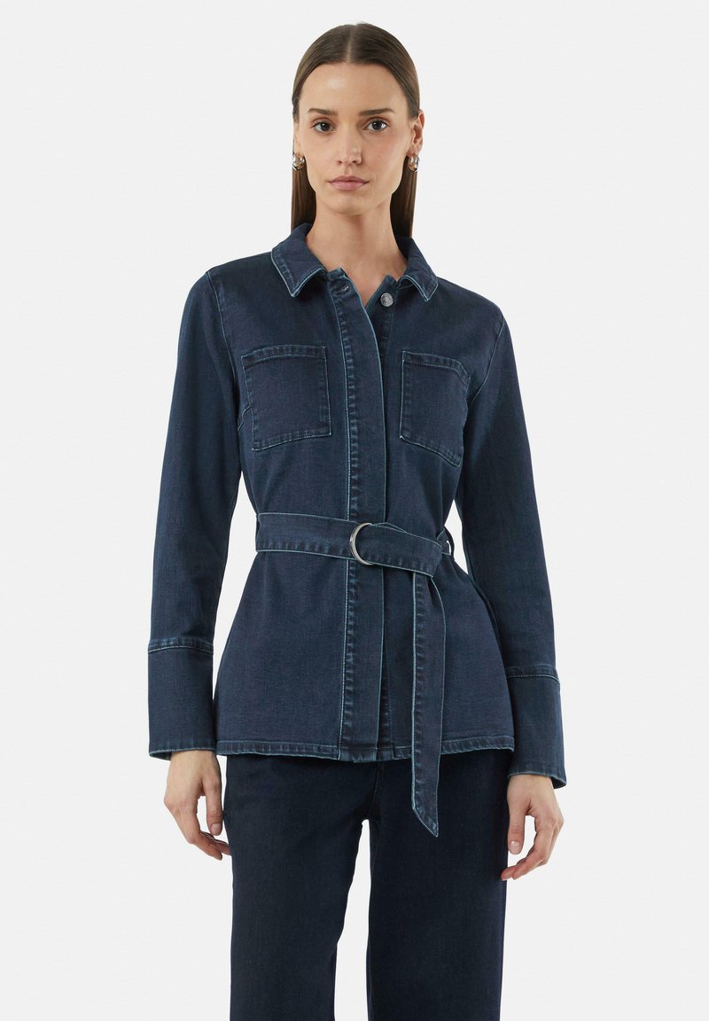 Donkerdenim jas met een kraag, voorzakken en een ceintuur in de taille. Beschikt over lange mouwen met manchetdetails en zichtbare stitching.