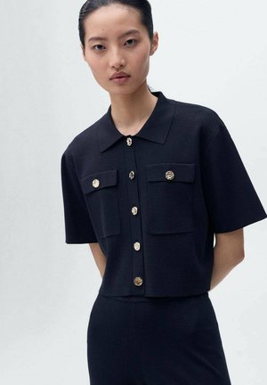 Top negro de manga corta con cuello tipo camisa, dos bolsillos frontales y botones dorados con un diseño texturizado. Tejido suave con un ajuste estructurado.