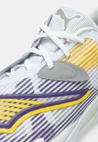 Detailní záběr na bílé tenisky Puma s fialovými a žlutými detaily, bílými tkaničkami a šedým semišovým patchem poblíž jazyka.