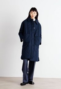 Manteau matelassé marine avec de grandes poches avant, une fermeture à boutons et un col. Porté sur un pantalon gris et des chaussures noires. Texture lisse.