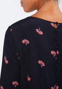 Marinblus med rosa blommönster, en nyckelhålsdetalj bak och knappdekorationer längs ryggen. Mjuk, lättviktig tyg.