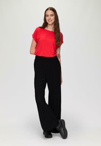 Rotes kurzärmliges Oberteil, kombiniert mit schwarzen, plissierten, weit geschnittenen Hosen. Das Model trägt schwarze Plateauschuhe und steht vor einem neutralen Hintergrund.