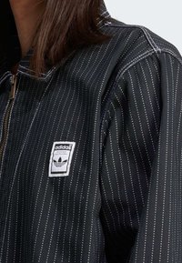 Černá bunda Adidas s vertikálními bílými tečkovanými pruhy, předním zipem a bílou nášivkou loga Adidas na levé straně hrudi.
