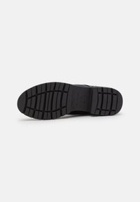 Suela de zapato negra con un patrón texturizado, que presenta surcos y crestas para mayor tracción. Incluye detalles del logotipo y bordes suaves.