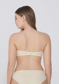 Béžová podprsenka bez ramínek se hladkým povrchem, s háčkovým zapínáním na zádech. Minimalistický design s širokým pásem a bez viditelných švů.