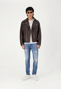 Smeđa kožna motociklistička jakna sa srebrnim detaljima, bijela majica, poderane plave traperice i bijele tenisice. Dodaci uključuju crne sunčane naočale.