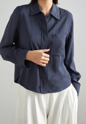 Persoon draagt een donkerblauw, geknoopt overhemd met borstzak, gecombineerd met losse witte broek, houdt het voorpand van het overhemd bij de knopen vast.