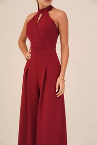 Combinaison bordeaux avec un col halter, présentant une découpe en forme de trou de serrure, des jambes larges plissées et un buste ajusté en tissu lisse. Détails en métal doré.