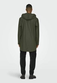 Cămașă lungă verde impermeabilă cu glugă, cu o croială dreaptă și două buzunare laterale, purtată peste pantaloni cargo negri și pantofi.