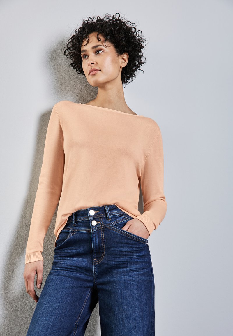 Pfirsichfarbener, langärmeliger Strickpullover mit weitem Halsausschnitt, kombiniert mit hoch geschnittenen Jeans aus Denim, die über zwei Knöpfe und seitliche Taschen verfügen.