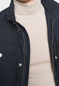 Béžový žebrovaný rolák nošený pod rozepnutou tmavě námořnickou bundou se stříbrnými patentkami a zipovými detaily.