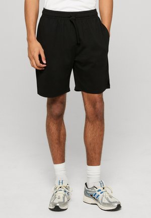 Mężczyzna w czarnych szortach z troczkiem, białych za kostkę skarpetkach oraz szarych i niebieskich butach sportowych, stojący na tle jednolitego szarego tła.