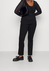 Top nero in pizzo a maniche lunghe con scollatura a zigzag, abbinato a pantaloni neri slim-fit e scarpe nere con dettaglio a fiocco. Inclusa anche una borsa nera con texture.
