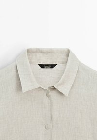 Chemise en lin beige clair avec col classique et deux boutons visibles à l'avant, dévoilant la partie supérieure de la poitrine et le décolleté.