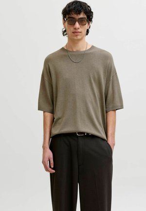 Ung mand iført brun, oversized kortærmet sweater, sorte bukser, sølved kædehalskæde og mørke rektangulære solbriller, stående med den ene hånd i lommen.