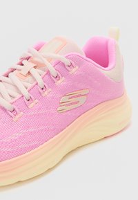 Zapatilla deportiva de color rosa claro con parte superior de malla texturizada, acentos en tonos pastel contrastantes y una suela acolchada. Presenta un detalle de logo dorado y cordones.
